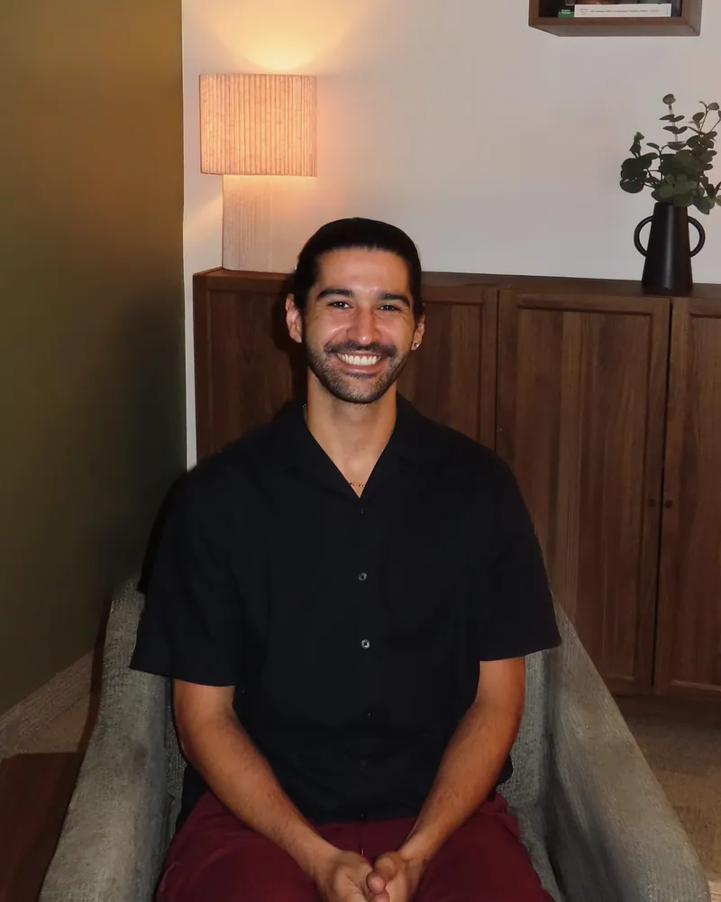 Sammy Pollino seated in a grey armchair against an olive wall, smiling